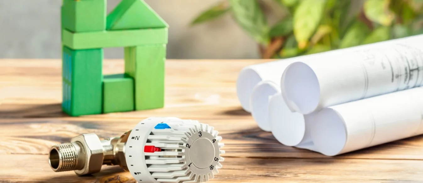 Haus aus grünen Bauklötzen, Thermostat eines Heizkörpers und Bauzeichnungen auf Holztisch, symbolisch für die Heizung mit erneuerbaren Energien.