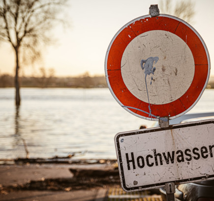 Durchfahrt-verboten-Schild und Schild mit der Aufschrift „Hochwasser“ vor überschwemmtem Flussufer.
