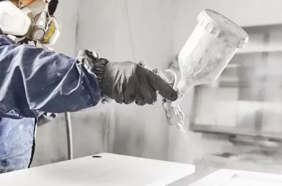 Mitarbeiter in Overall, mit Atemschutzmaske und Farbspritzpistole lackiert in Lackierkabine hölzerne Schranktüren.