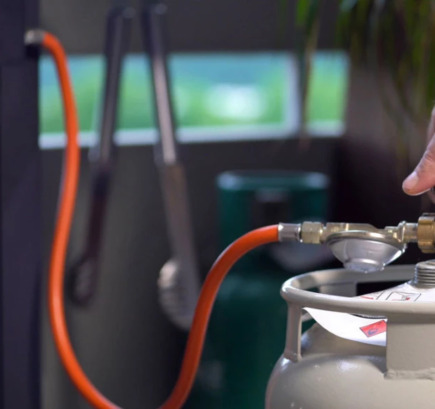 Hand, die den Hahn einer Propangasflasche aufdreht, im Hintergrund ein Gasgrill.